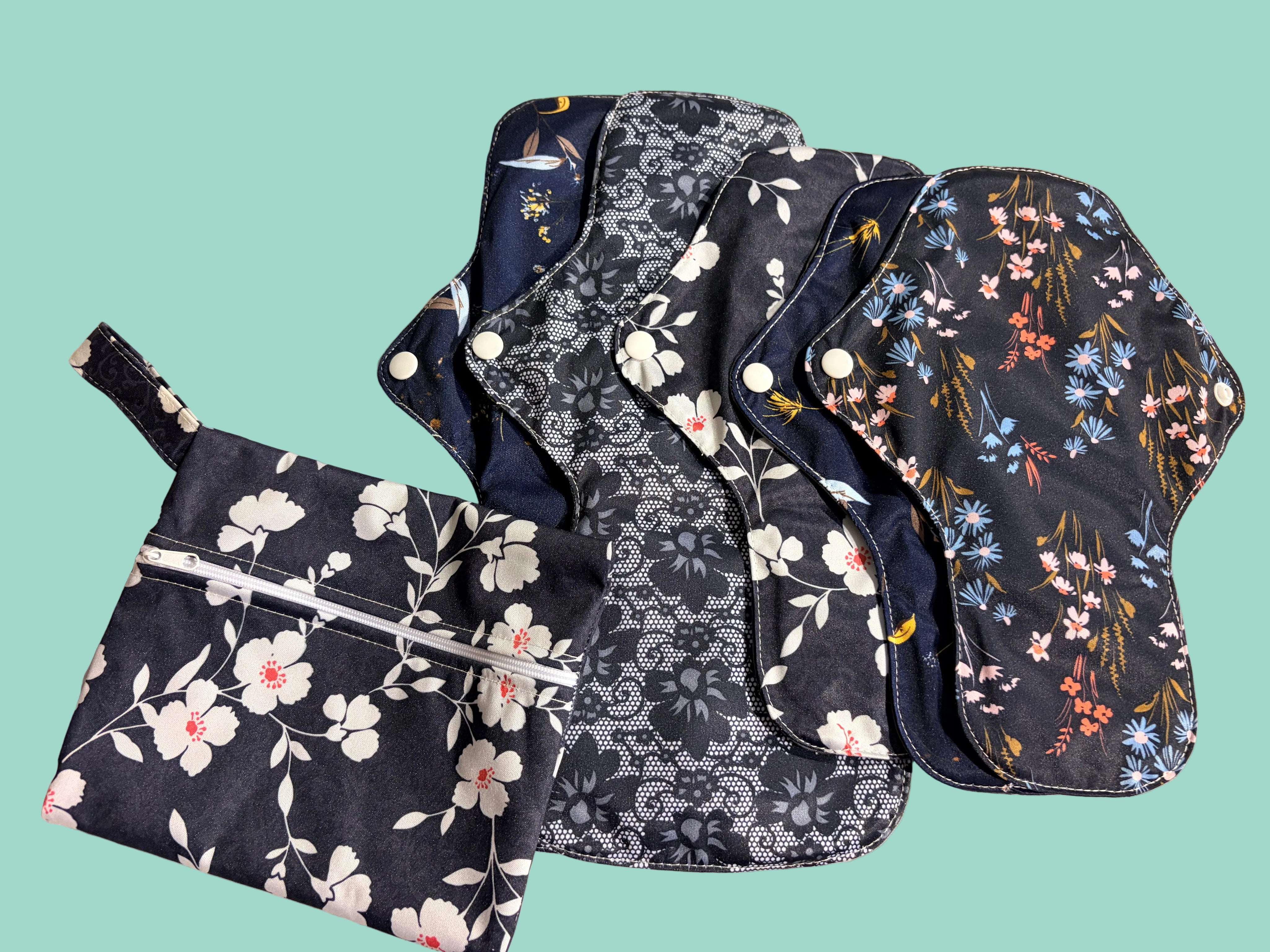 A set of reusable cloth pads and a matching zippered pouch laid out on a light fabric surface. The items feature dark navy backgrounds with white floral prints and multicolored floral motifs in pink, blue, yellow, and orange.