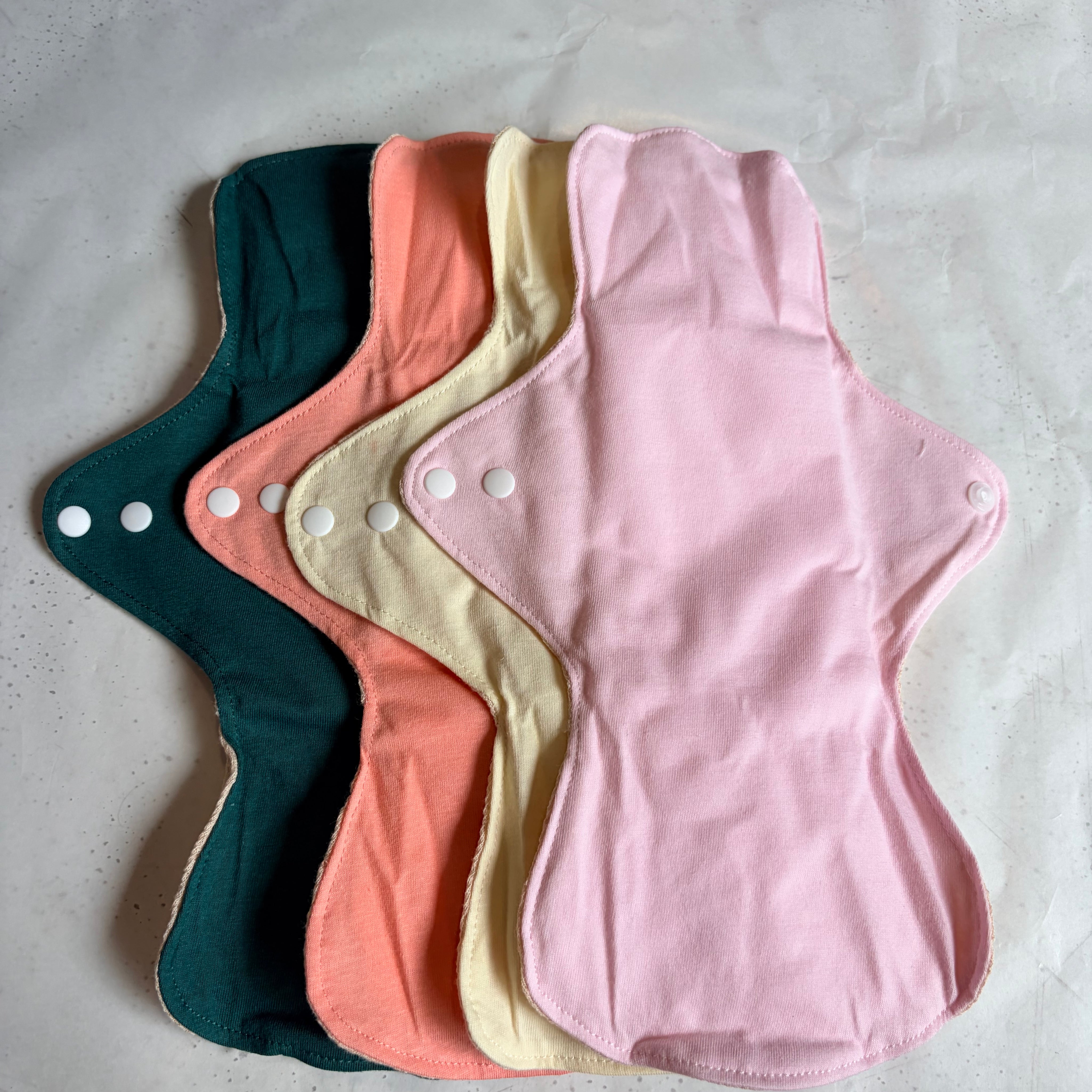 A stack of reusable cloth menstrual pads in pastel colors, including pink, cream, coral, and dark teal, arranged on a light textured surface.